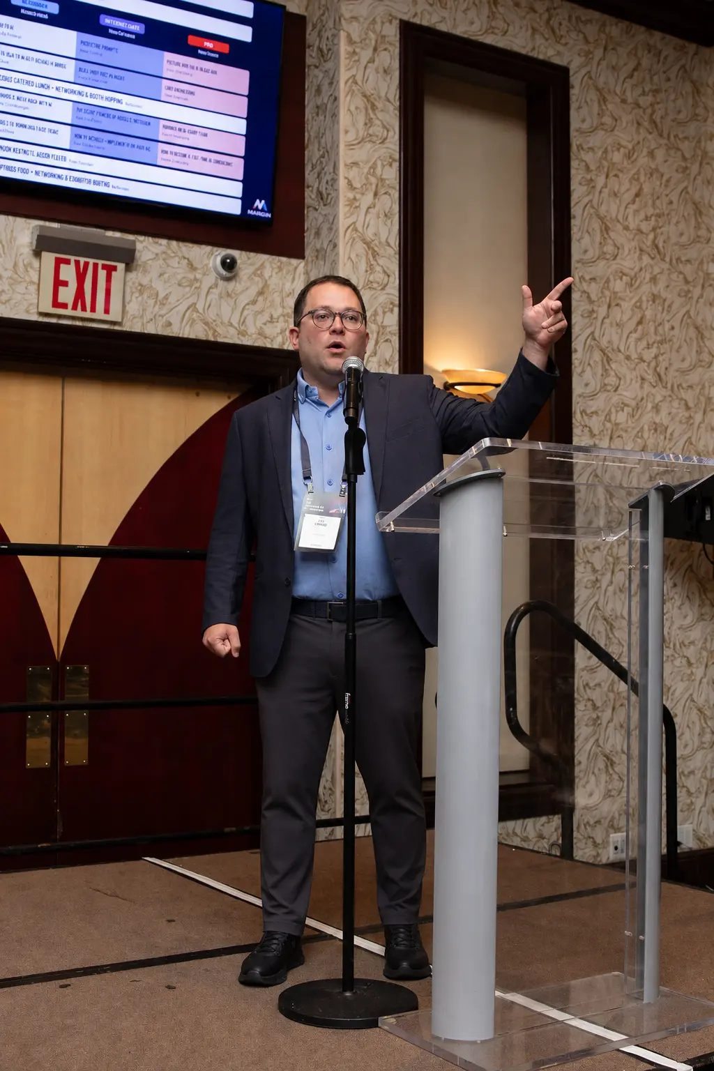Eli Langer speaking at the podium during The Practical AI Conference, session schedule on screen behind him